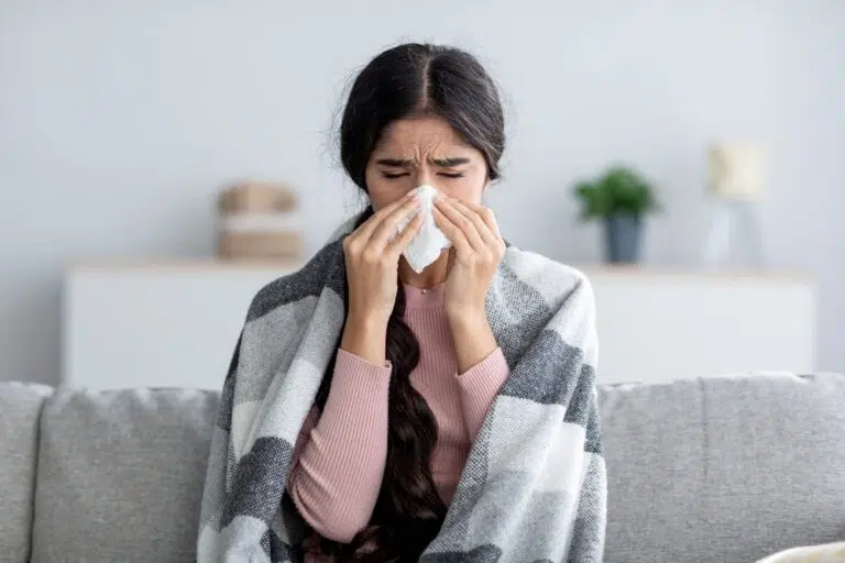 A woman with dark brown hair and a pink shirt sits on a couch. She is wrapped in a blanket and holds a tissue to her nose.