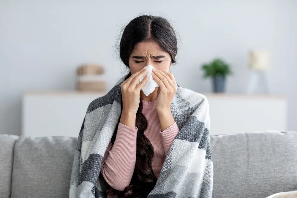A woman with dark brown hair and a pink shirt sits on a couch. She is wrapped in a blanket and holds a tissue to her nose.