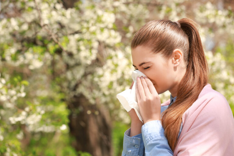 A woman in a denim shirt with brown hair stands and blows her nose into a tissue. In the background, a tree is full of white blossoms.